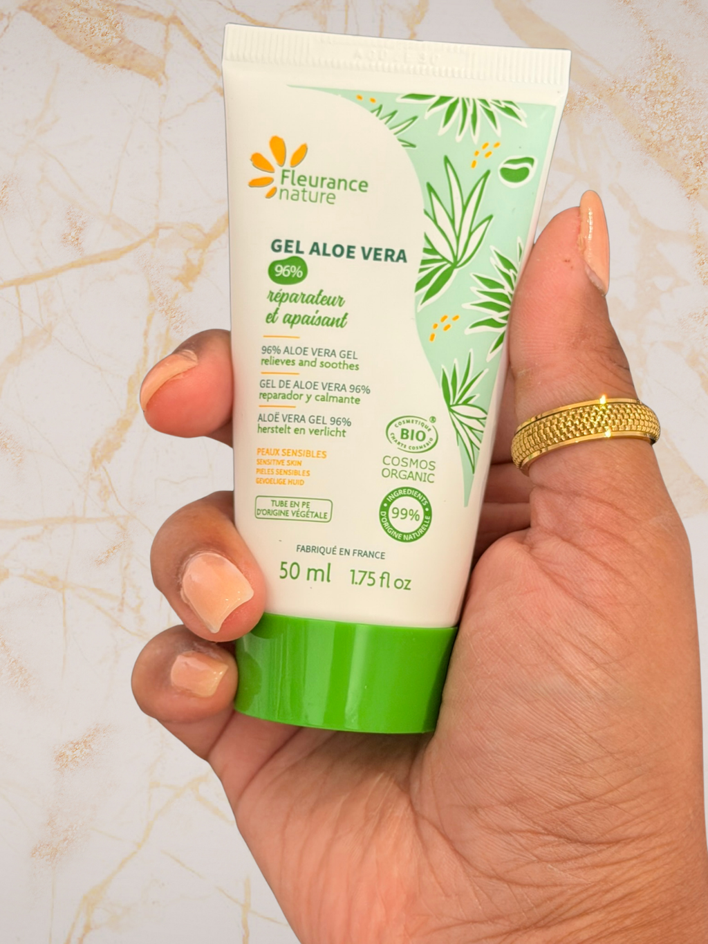 A hand holding a 50 ml squeeze tube of Fleurure nature Gel Aloe Vera hand and skin repair gel, set against a light beige marble background.